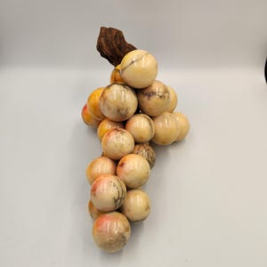 May include: A decorative cluster of grapes made from polished stone. The grapes are a mix of cream, peach, and brown tones, with a wooden stem. The grapes are round and smooth, and the wooden stem is textured.