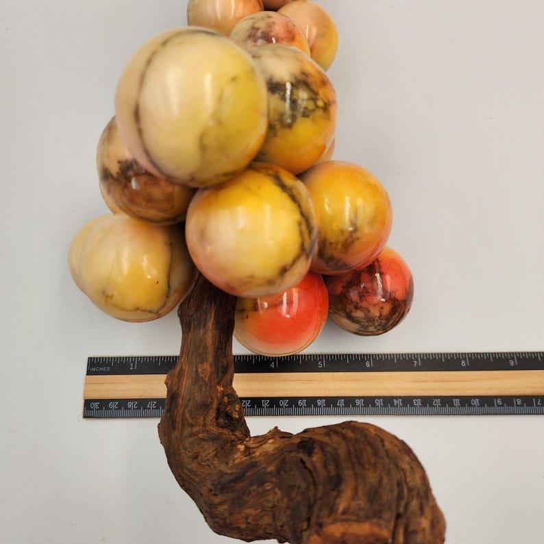 May include: A decorative sculpture of a cluster of marble spheres in shades of yellow, orange, and red, attached to a natural wood base. The spheres vary in size and have unique veining patterns. A ruler is placed next to the sculpture.