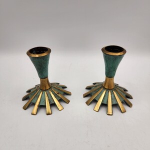 May include: A pair of metal candle holders featuring a flared, trumpet-shaped top and a wide, sunburst-style base. The design combines a polished gold-tone finish on the base rays and central stem with a textured, oxidized teal-green patina.