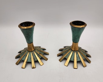 May include: A pair of metal candle holders featuring a flared, trumpet-shaped top and a wide, sunburst-style base. The design combines a polished gold-tone finish on the base rays and central stem with a textured, oxidized teal-green patina.