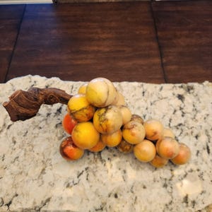 May include: A decorative cluster of grapes made from polished stone. The grapes are a mix of yellow, orange, and reddish-brown hues, with a wooden vine stem. The grapes are arranged on a speckled countertop.