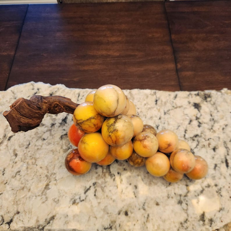 May include: A decorative cluster of grapes made from polished stone. The grapes are a mix of yellow, orange, and reddish-brown hues, with a wooden vine stem. The grapes are arranged on a speckled countertop.