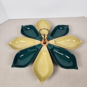 May include: A flower-shaped serving dish with eight petal-shaped compartments. The dish alternates between teal and yellow-cream colors. A brass handle rises from the center. The dish is likely made of ceramic or porcelain.