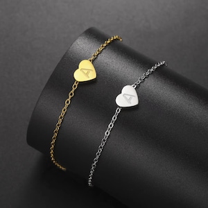 May include: Two delicate bracelets, one gold-toned and one silver-toned, each featuring a heart-shaped charm. The charms are engraved with the letter "A". The bracelets are displayed on a dark, textured surface.