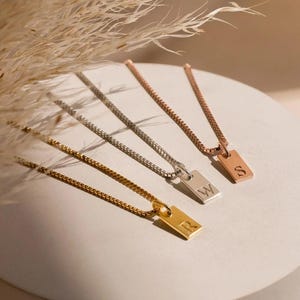 Può includere: Quattro collane con ciondoli rettangolari, ognuno con un'iniziale. I ciondoli sono in oro, argento e oro rosa. Le catene sono sottili e i ciondoli piccoli, adatti per essere sovrapposti. Presentate su una superficie neutra.