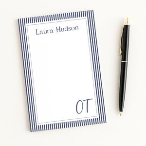 May include: A rectangular notepad featuring a white center with a dark blue vertical striped border. The name Laura Hudson is printed at the top, and the letters OT are printed in the bottom right corner. A black ballpoint pen with gold accents.