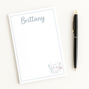 May include: A white notepad with a light blue border and the name "Brittany" in script. A cartoon tooth with dental tools is in the corner. A black pen with gold accents rests on the side.
