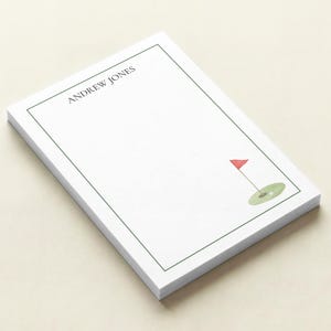 May include: A stack of white notepads with a green border and a golf-themed illustration. The illustration includes a red flag, a green putting green, and a golf ball. The text "ANDREW JONES" is printed at the top.