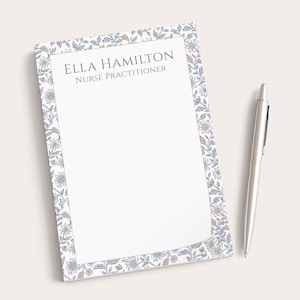 May include: A white notepad with a floral border in blue, tan, and gray, with the text "ELLA HAMILTON NURSE PRACTITIONER." A silver pen rests on the right side of the notepad.