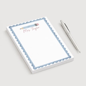 May include: A rectangular notepad with a scalloped blue border, featuring a watercolor illustration of a pencil and the text Mrs. Taylor in pink script. A silver metal ballpoint pen is positioned to the right of the notepad on a plain surface.