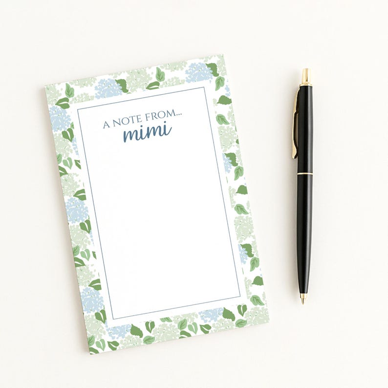 May include: A notepad with a floral design featuring blue and green hydrangeas, with the text "A note from... Mimi." A black pen with gold accents rests beside the notepad on a white surface.