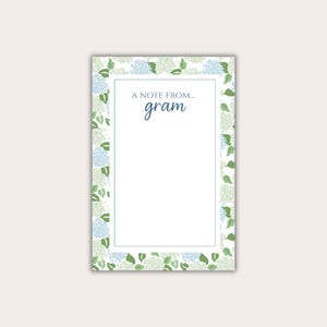 May include: A rectangular notepad with a white interior and a floral border. The border features light blue and green hydrangea blooms and leaves on a white background. The text "A NOTE FROM... gram" is printed at the top.
