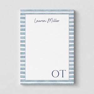 May include: A rectangular notepad featuring a white center bordered by horizontal blue and white stripes. The top section displays the name Lauren Miller in dark blue script, while the bottom right corner features the dark blue capital letters OT.