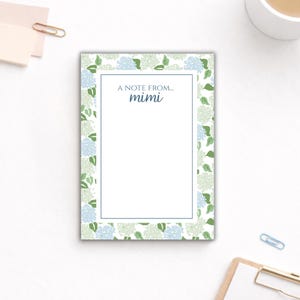 May include: A rectangular notepad featuring a floral hydrangea pattern in blue and green. The notepad has a white interior with the text "A note from... mimi" in blue. A blue border frames the white interior.