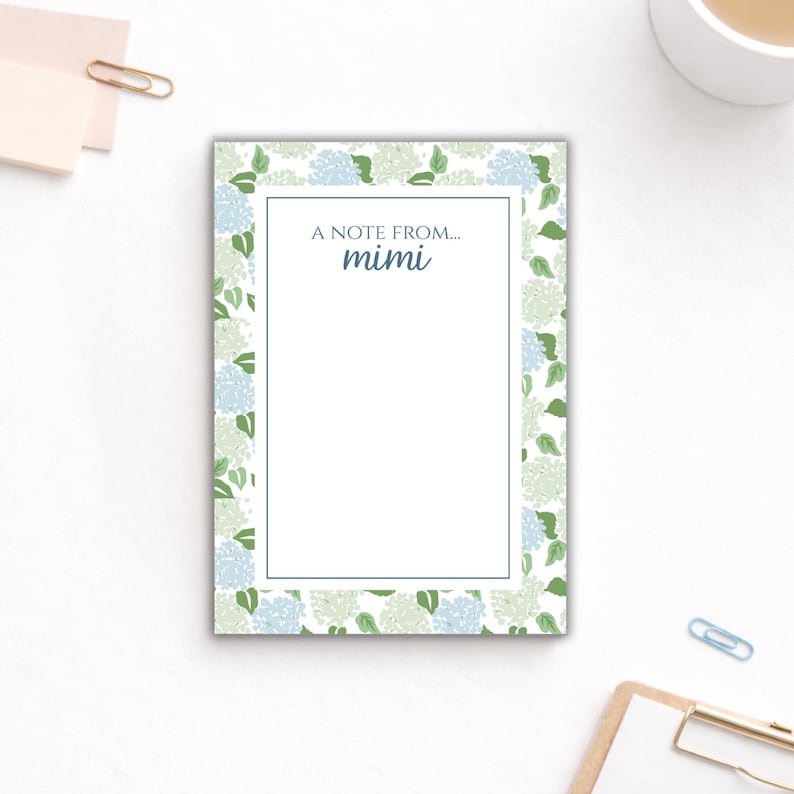 May include: A rectangular notepad featuring a floral hydrangea pattern in blue and green. The notepad has a white interior with the text "A note from... mimi" in blue. A blue border frames the white interior.