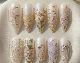 Könnte beinhalten: Ein Set aus zehn mandelförmigen, künstlichen Nägeln mit Perlmutt-Effekt. Jeder Nagel zeigt aufwendige Designs in Gold, Rosa und Grün, darunter ein Mond, Blumenmuster und ein Schwan. Die Nägel sind auf einer weißen Oberfläche angeordnet.