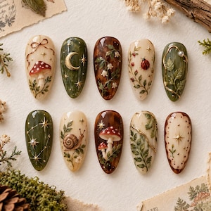 May include: A set of ten almond-shaped press-on nails featuring intricate designs including red mushrooms, a gold crescent moon, lily of the valley, a ladybug, green leaves, a snail, and gold star accents on cream, olive green, and brown backgrounds.