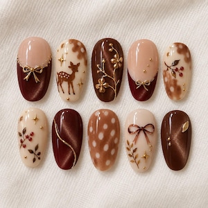 May include: A set of ten press-on nails featuring a brown and cream color palette. Designs include a deer silhouette, gold bows, floral branches, red berries, white polka dots, and metallic accents on glossy, cat-eye, and solid finish surfaces.
