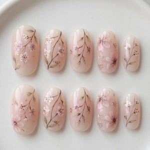 May include: A set of ten oval-shaped press-on nails featuring a soft nude base colour. Each nail is decorated with delicate hand-painted floral patterns in shades of pink and white, accented with thin green stems and small gold foil flecks.