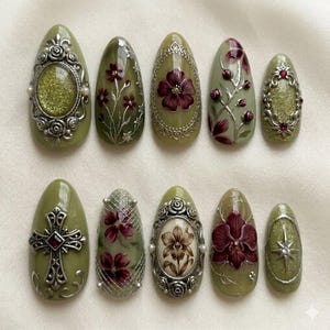 May include: A set of ten olive green artificial nails with intricate designs. Some nails feature silver frames with glitter, floral patterns, or a cross. Others showcase detailed flower paintings in shades of burgundy and brown.