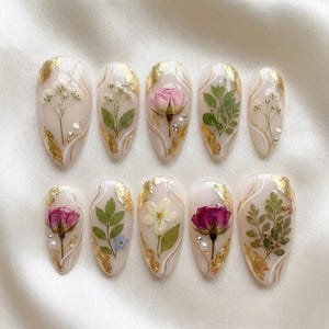 May include: A collection of ten almond-shaped artificial nails, each decorated with dried flowers, gold leaf, and tiny pearl details. The nails showcase diverse floral arrangements, including roses and delicate white flowers, set on a clear base.