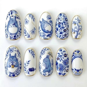 May include: A set of ten white and blue nail art designs. Each nail features a different design, including floral patterns and cartoon characters. The nails have a gold trim.