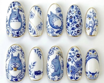 Könnte beinhalten: Ein Set aus zehn weißen und blauen Nageldesigns. Jeder Nagel hat ein anderes Design, darunter Blumenmuster und Zeichentrickfiguren. Die Nägel haben einen goldenen Rand.