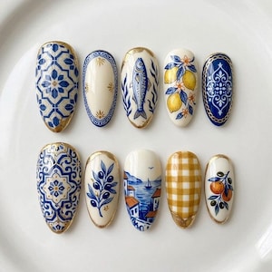 Könnte beinhalten: Ein Set aus zehn mandelförmigen Nageldesigns. Jeder Nagel zeigt ein einzigartiges handgemaltes Design, darunter blau-weiße Fliesenmuster, ein Fisch, Zitronen, Oliven, eine Küstenlandschaft und ein Vichy-Muster. Goldfarbene Verzierungen akzentuieren jeden Nagel.