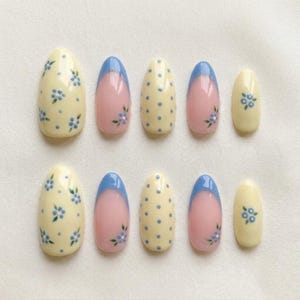 Pode incluir: Um conjunto de dez unhas postiças em formato amendoado, com uma mistura de designs incluindo amarelo pálido com padrões florais azuis, rosa com pontas francesas azuis e pequenas flores, e amarelo pálido com bolinhas azuis brilhantes.