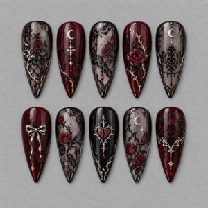 May include: A set of ten almond-shaped press-on nails featuring deep red and black color palettes. Designs include intricate black lace patterns, red roses, silver metallic filigree, crescent moons, a silver bow, and a broken heart emblem.