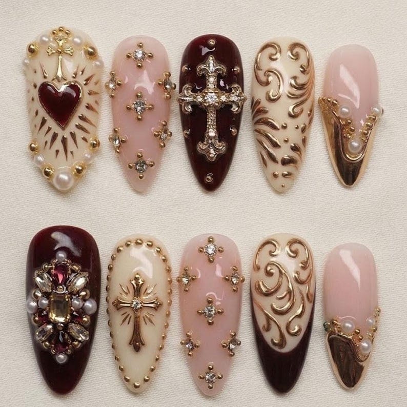 May include: A set of ten artificial nails in various designs. The nails feature a range of colors, including burgundy, cream, and pink, and are adorned with gold embellishments, rhinestones, and pearl accents. Some nails showcase cross and heart motifs.
