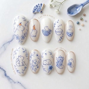 May include: White nail art with blue capybara illustrations, flowers, and teacups. The designs include the text "EASY MIX & MATCH" and "CLEAN GIRLS VIBE". The nail art is elegant and cute, with a clean aesthetic.