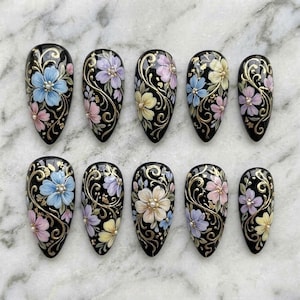 May include: A set of ten almond-shaped press-on nails with a black base, featuring hand-painted floral designs in shades of blue, pink, yellow, and lavender, accented with gold swirling filigree patterns and small white center beads on each flower.