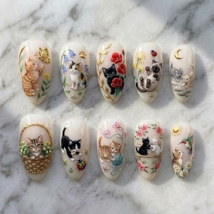 Peut inclure: Un ensemble de dix faux ongles en amande ornés d'illustrations peintes à la main de chatons dans diverses postures, agrémentés d'éléments en feuille d'or, de fleurs, de papillons, de coccinelles, d'un panier tressé et de symboles célestes.