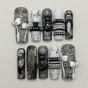 Könnte beinhalten: Ein Set aus zehn langen, aufklebbaren Nägeln mit Musik- und Katzenmotiv. Designs umfassen Noten, Violinschlüssel und schwarze Katzenfiguren. Die Farben sind Schwarz, Weiß und Grau, mit silbernen Akzenten und Nietendetails.