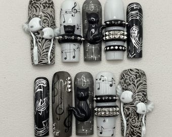 May include: A set of ten long, press-on nails with a music and cat theme. Designs include musical notes, treble clefs, and black cat figurines. Colors are black, white, and gray, with silver accents and stud details. Some nails feature 3D elements.