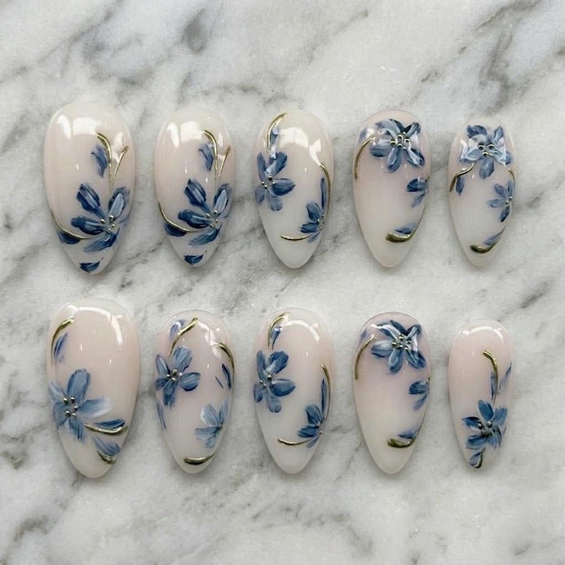 Puede incluir: Un conjunto de diez u&ntilde;as postizas de forma ovalada. Cada u&ntilde;a est&aacute; pintada con una base rosa claro y presenta un dise&ntilde;o floral azul con detalles dorados. Las u&ntilde;as est&aacute;n dispuestas sobre una superficie de m&aacute;rmol blanco.