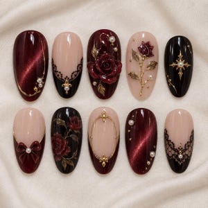 May include: A set of ten almond-shaped press-on nails featuring a mix of deep burgundy cat-eye polish, nude bases with black lace patterns, 3D red rose embellishments, gold metallic accents, small white pearls, and intricate black bow designs.