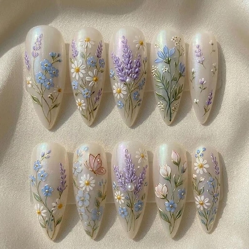 May include: A set of ten almond-shaped artificial nails, each adorned with delicate floral designs. The nails feature a pearlescent base with hand-painted details of lavender, daisies, and blue flowers, some with a butterfly accent.