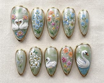 May include: A set of ten almond-shaped press-on nails featuring hand-painted designs on a sage green background. The artwork includes white swans on water, blue forget-me-nots, orange and pink wildflowers, white daisies, and lily flowers.