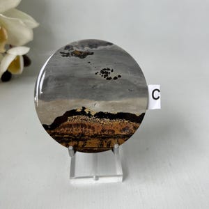 May include: A round, polished stone with a landscape design. The stone features a mix of gray, brown, and tan colors, resembling a natural scene. The stone is displayed on a clear stand, with a small white tag labeled "C".