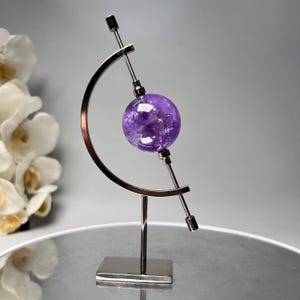 以下が含まれることがあります： 約7.6cmの直径の研磨されたアメジストの球体が、モダンなシルバー調の金属製スタンドに保持されています。スタンドは湾曲したアームと正方形のベースを備え、宝石のミニマリストなディスプレイを作成しています。