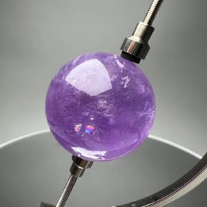 Peut inclure: Une sphère de cristal d'améthyste polie, de couleur violet profond. La sphère présente des structures cristallines internes et une surface réfléchissante. Montée sur un support métallique avec un col flexible.