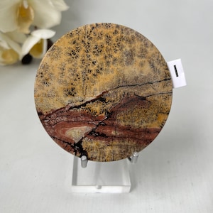 May include: A circular, polished stone with a distinctive pattern. The upper section displays a tan and black speckled design, while the lower half has reddish-brown and black veining. The stone is presented on a clear stand.