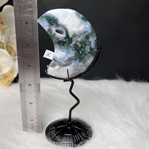May include: A crescent-shaped, polished stone with green, white, and blue patterns, mounted on a black metal stand. The stone is approximately 5 inches in height, and the stand has a decorative base.