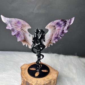 May include: A black metal sculpture of two figures embracing, with amethyst wings. The sculpture is mounted on a wooden base. The wings are a mix of purple, white, and brown. The figures have horns and tails.