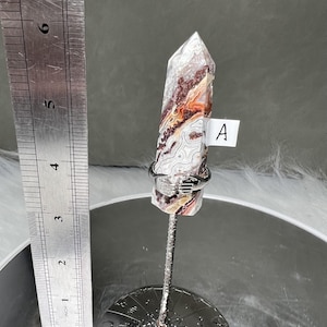 Puede incluir: Una punta de cristal con una superficie estampada en tonos blancos, marrones y rojos, sujeta por una mano de metal plateado. El cristal mide aproximadamente 13 cm de largo. Una pequeña etiqueta blanca con la letra "A" está adjunta.