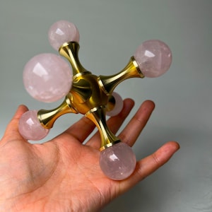 Può includere: Un oggetto decorativo con una struttura metallica color ottone e sei pietre sferiche di quarzo rosa. L'oggetto è tenuto in mano, mostrando il metallo lucido e la qualità traslucida delle pietre rosa.
