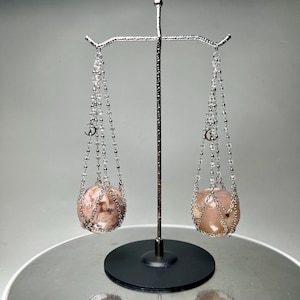 Pode incluir: Uma balança decorativa com duas esferas rosa-acastanhadas suspensas por correntes. As correntes são adornadas com contas transparentes e pequenos amuletos prateados. A balança é suportada por uma base preta e um braço de metal prateado.
