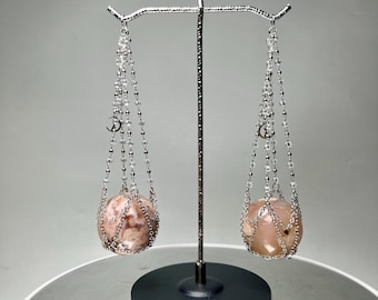 May include: A decorative balance scale with two pinkish-tan spheres suspended by chains. The chains are adorned with clear beads and small silver charms. The scale is supported by a black base and a silver metal arm.
